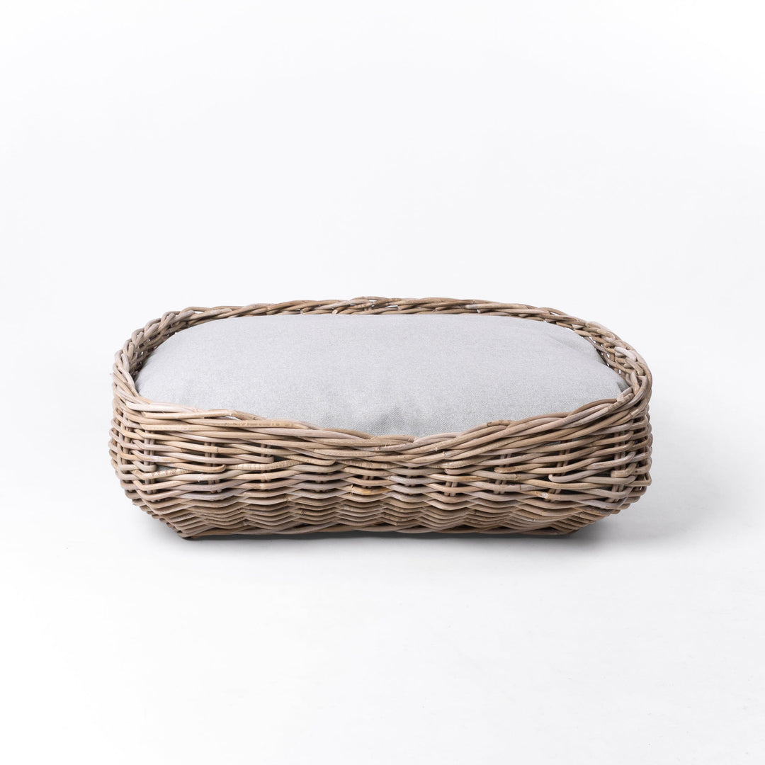A rattan dog basket with a pale beige cushion, on a white background.
