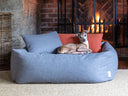 Fawn coloured Italian Greyhund d go lying in a grey luxury dog bed with two small cushions in the bed in grey and orange, in front of a fireplace on a natural hessian rug.