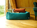 An Italian Greyhound Dog lying in a jade colour bolster dog bed with two mini cushions in orange and lemongrass in a modern house with a view to the outdoors.
