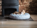 A dog lying on a round pale grey dog bed mattress in front of a wood burning stove in a house with a stone wall backdrop and wooden floor