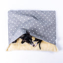 Seen from above, a black dog peeking out of a Charley Chau luxury dog burrow bag, a dog sleeping bag dog bed, in a pale grey polka dot fabric with a cream faux-fur lining on a white backdrop.