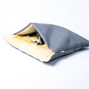 A grey luxury dog burrow bag, a sleeping bag style dog bed, with a cosy cream Faux-Fur lining with a dog sleeping very cosy inside on a white backdrop.