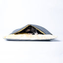 A dog lying inside a grey luxury dog burrow bag, a snuggle sack type of dog bed, with a cosy cream Faux-Fur lining on a white backdrop.
