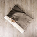 Overhead view of a dog peeking out of a luxury Dog Burrow Bag with brown velvet outer cover and plush cream faux-fur lining interior in a home setting with a wooden floor.