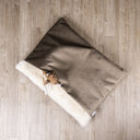 Overhead view of a dog peeking out of a luxury Dog Burrow Bag with brown velvet outer cover and plush cream faux-fur lining interior in a home setting with a wooden floor.
