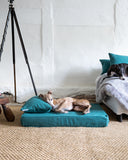 Dog lying on a jade green memeory foam mattress style dog bed in a room with a white wall and hessian floor. 
