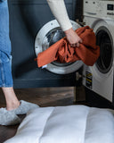 Person putting an orange dog bed cover into a washing machine in a laundry room.