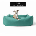 A fawn dog lying in a luxury box dog bed with high sides - The Bliss Bolster Bed - by designers Charley Chau, on a white background with a press feature badge for "Elle Decoration" above it.