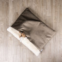 A luxurious sleeping bag style dog bed made with a brown velour and a cosy cream faux-fur fleece lining with the head and paws of a fawn and white coloured Italian Greyhound dog peeping out on an oak wooden floor.