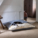 An Italian Greyhound dog sleeping very cosy inside a grey luxury cave bed style Dog Snuggle Bed with a plush cream faux-fur fleece lining in a home setting with a natural hessian floor and white  walls.