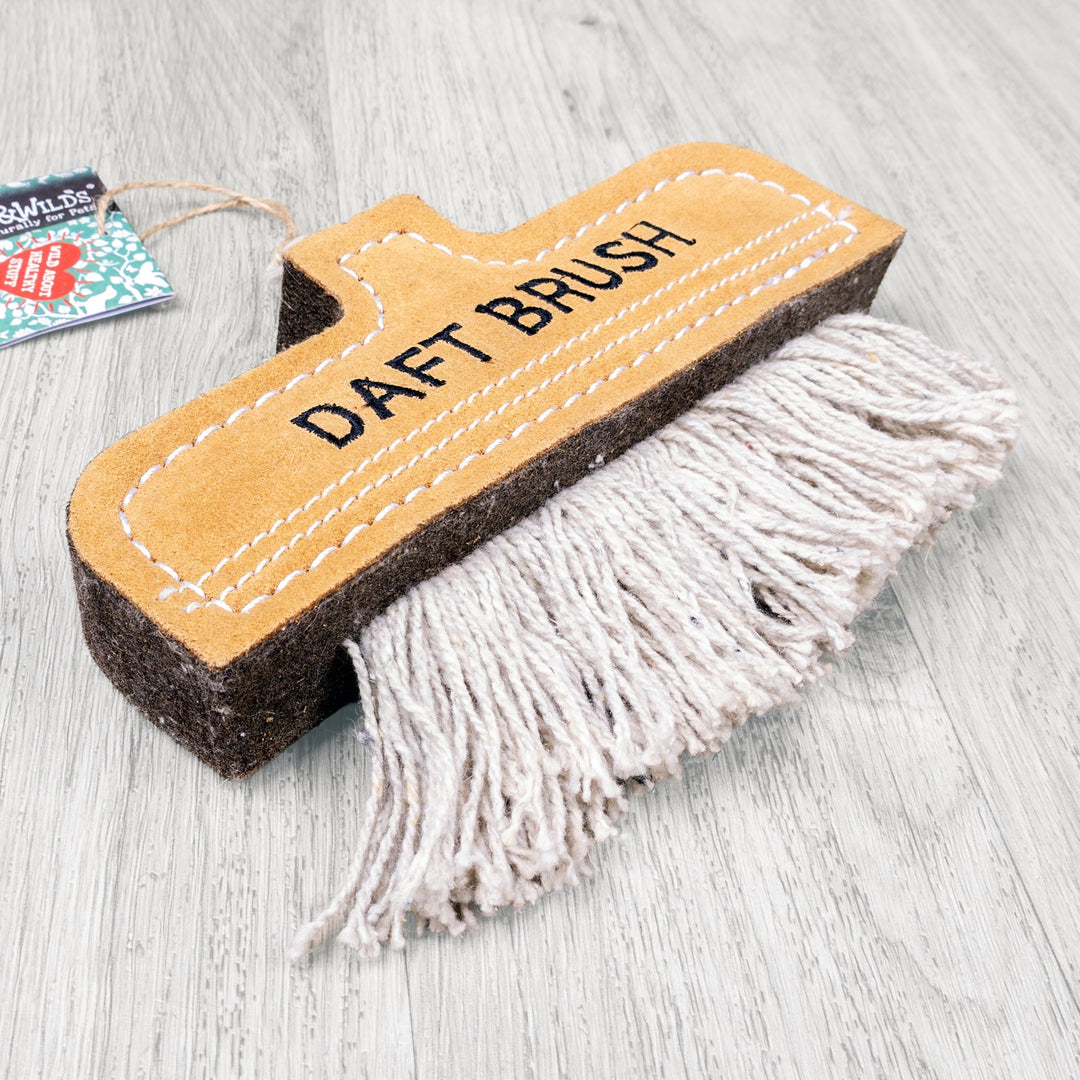 An eco-friendly dog toy in the shape of a cleaning brush in a tan coloured suede with grey string on a pale grey oak floor.
