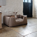 Dog lying in a premium quality high-sided box dog bed in a beige colour in a room with stone flooring, white walls  and a dark green door.