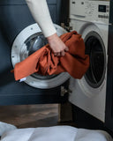 A person's hand and arm are shown loading an orange terracotta-colored dog bed cover from a machine washable dog bed into a white front-loading washing machine, with a dark cabinet behind it.