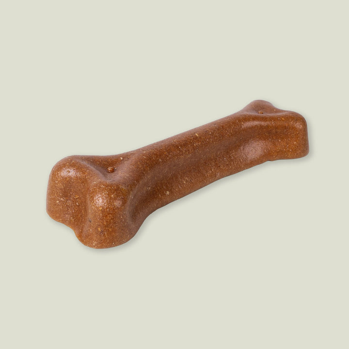 A brown, bone-shaped, natural dog chew treat made from Madeira Cheese and Peanut Butter on a pale sage green background.