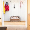 Italian Greyhound dog lying on a luxury  Raised Rattan Dog Bed placed in  hallway with some coats and dog leads hanging on the wall above the raised rattan dog bed.