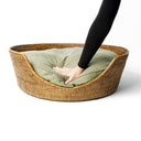 A golden tan colour round Rattan Dog Basket with a fern green dog bed mattress inside a person's hand pressing down on the luxurious mattress 