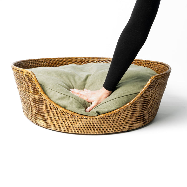 A golden tan colour round Rattan Dog Basket with a fern green dog bed mattress inside a person's hand pressing down on the luxurious mattress 