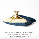 A small dog lying inside a luxury dog snuggle bed, a cave bed style dog bed or sleeping bag style dog bed, with a dark navy blue Velour outer cover and plush, cream faux-fur lining on a white backdrop.