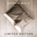 A luxury sleeping bag style dog bed made with a velour in mid-brown and lined with a cream faux-fur fleece lining with the head of a fawn and white dog peeping out and placed on an oak floor.