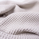 A close-up of a pale beige cotton waffle blanket with a mini-waffle texture, showing its textured grid pattern and soft folds.
