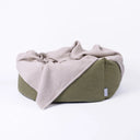 An olive green luxury donut dog bed with a light beige waffle texture cotton dog blanket draped in the bed and over the front, on a plain white studio background.