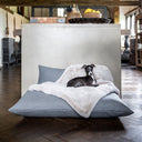 Dog lying on a very plush, stylish pale grey faux-fur dog blanket on top of a deep-filled luxury dog bed mattress in a home setting with dark wooden floor.