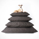 A dog lying on top of a stack of four different size luxury dog bed mattresses in a dark brown/black woven upholstery fabric on a white backdrop.