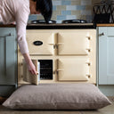 person opening the oven door of a cream coloured Aga range cooker with a luxury deep-filled dog bed mattress on the floor in front of the range cooker.