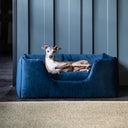 A dog lying in a luxurious, deep-sided navy blue velvet dog bed in a home setting with a natural carpet on a wooden floor. 