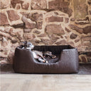 A dark brown high-sided Deeply Dishy Dog Bed by designers Charley Chau with an Italian Greyhound dog lying in the bed looking calm and content, in a home setting with a stone wall backdrop and stone and wood floor.