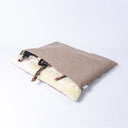 A black dog sleeping covered up in a snuggle bag style Dog Burrow Bag with a beige woven upholstery fabric outer cover and plush cream faux-fur lining on a white backdrop.