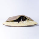A black dog sleeping covered up in a snuggle sack style Dog Burrow Bag with a beige woven upholstery fabric outer cover and plush cream faux-fur lining on a white backdrop.