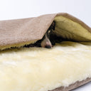 Close up of a black dog sleeping covered up in a snuggle bag style Dog Burrow Bag with a beige woven upholstery fabric outer cover and plush cream faux-fur lining on a white backdrop.