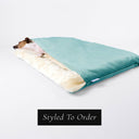 A dog sleeping very cosy in a bespoke Dog Burrow Bag with a pale turquoise velvet outer cover and plush cream faux-fur lining on a white backdrop.