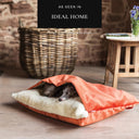 A dog tucked up cosy in a luxury dog snuggle bed with a built-in cover in orange velvet outer cover and cream faux-fur fleece lining in a home setting with a rattan log basket and wooden side table, with an 'As seen in Ideal Home' press feature badge above.