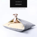Fawn Italian Greyhound nestled in grey dog cave snuggle bed with soft cream interior on a white backdrop and 'As seen in Stylist" press feature badge above.