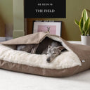 A gray Whippet dog sleeping inside a cozy, dog snuggle bed, a cave bed style dog bed, made with a woven textured upholstery-quality outer fabric cover and lined with a plush, cream faux-fur fleece lining. The bed is placed on a light-colored carpet in a home setting with an olive green wall, and a framed picture of a Whippet in the background.