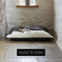 A small fawn dog sleeping cosy inside a luxurious Dog Snuggle Bed, a sleeping bag / cave bed style dog bed, with a dark brown outer cover and cream plush lining, in a home settnig on a stone floor with a whitewashed stone wall in the background.