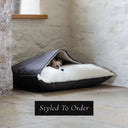 A small fawn dog sleeping cosy inside a luxurious Dog Snuggle Bed, a sleeping bag / cave bed style dog bed, with a dark brown outer cover and cream plush lining, in a home settnig on a stone floor with a whitewashed stone wall in the background.