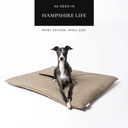 A black dog lying on a beige luxury dog travel bed mattress on a white backdrop with a featured in the press badge 'As seen in Hampshire Life' above.