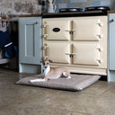 A dog lying on a beige colour luxury dog travel pad mattress ina durable, hard-wearing upholstery fabric, in a home setting with a cream Aga range cooker in the background on a stone floor.