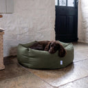 An olive green, water-resistant donut dog bed with a brown working cocker spaniel dog sleeping very comfortably inside. The water-resistant dog bed has high sides in the bolster nest and is set in a country-style home on a stone floor against a white-washed stone wall and a dark wooden door.