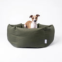 A water-resistant donut style dog bed by Charley Chau, with high bolster sides, made from a tough, water-resistant Dry Wax Cotton Canvas in olive green, with a small brown and white dog sitting inside, on a white background.