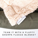 A close-up of a beige sherpa fleece blanket draped over a dark olive green, water-resistant donut dog bed, with a visible sewn label reading 'Charley Chau' and text at the bottom that says 'TEAM IT WITH OUR SHERPA FLEECE BLANKET.'