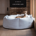 A dog lying in a luxury dog donut bed with high sides in pale grey, with a cream faxu-fur blanket in a home setting with "As seen in Country Life" press feature badge.