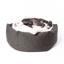A black dog sleeping in a high-sided, calming donut dog bed made with a dark brown/black woven upholstery fabric and grey sherpa fleece dog blanket on a white backdrop.