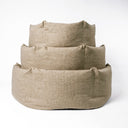 A stack of three high-sided, calming donut dog beds in three different sizes made with a beige woven upholstery fabric on a white backdrop.