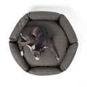 A dog lying in a high-sided, calming dog donut bed made with a dark brown/black woven upholstery fabric seen from above on a white backdrop.