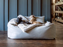 Two dogs  sleeping soundly in a pale grey luxury dog bed made to last, looking very cosy on pale grey a faux-fur blanket, in a home setting with a dark wooden parquet floor and a blue wood panelled wall behind the dog bed. 