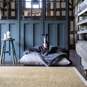 A black dog sitting on a plush machine washable faux-fur dog blanket in a grey blue colour called Russian Blue ona a luxury dog bed mattress in a home setting with a deepl blue grey wooden panelled wall and natural hessian rug.